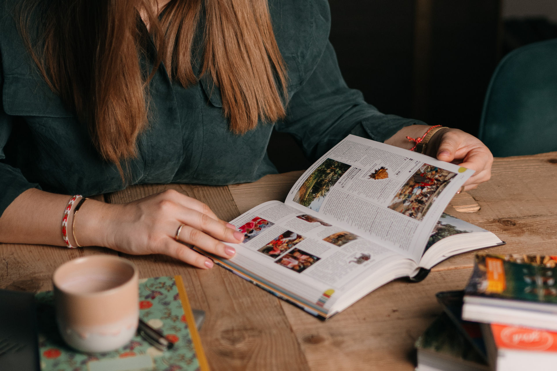Frau blättert in einem aufgeschlagenen Buch und sitzt an einem Holztisch; eine Tasse steht daneben.
