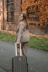 Frau im Hosenanzug zieht einen Rollkoffer hinter sich her und blickt lächelnd über die Schulter, während sie an einer Backsteinwand mit Herbstlaub vorbeigeht.