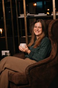 Frau sitzt entspannt in einem Sessel, hält eine Tasse in den Händen und lächelt in die Kamera.