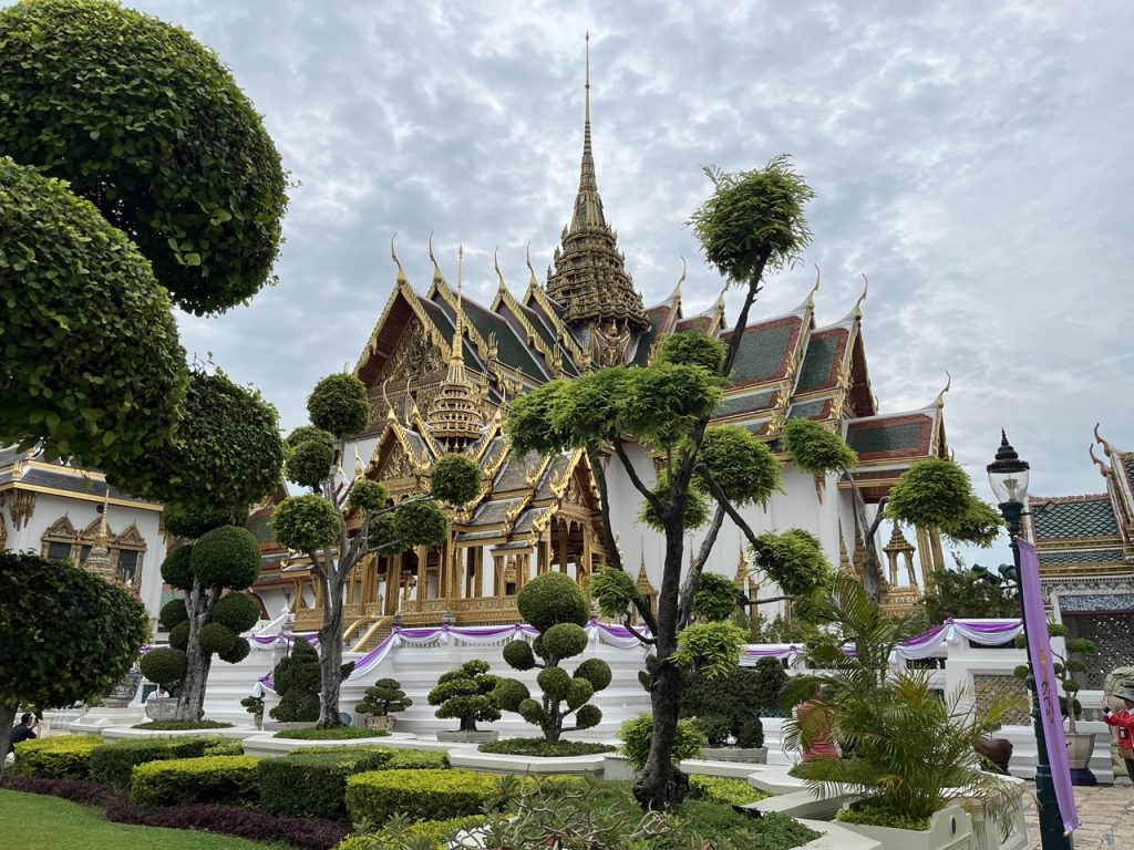 Prunkvoller Tempel in Bangkok mit goldenen Verzierungen und spitzen Dachgiebeln, umgeben von sorgfältig getrimmten Bäumen und einer gepflegten Gartenanlage.