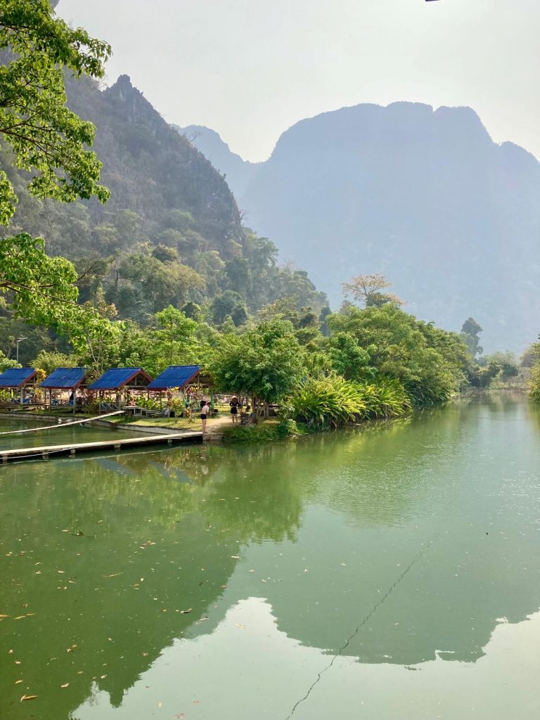 Ruhiger See inmitten einer grünen Berglandschaft; am Ufer stehen kleine Hütten mit blauen Dächern, und die umliegenden Bäume spiegeln sich im Wasser.